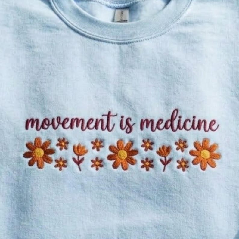 K&ouml;nnte beinhalten: Hellblaues Sweatshirt mit dem gestickten Schriftzug "movement is medicine" in Burgunderrot. Unter dem Text befindet sich eine Reihe gestickter orangefarbener und gelber Blumen, die ein Blumenmuster bilden.