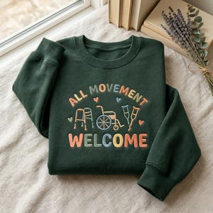 May include: Dark green sweatshirt with the words "ALL MOVEMENT WELCOME" in colorful, embroidered letters. The design includes illustrations of a walker, wheelchair, and crutches, with small heart accents. The sweatshirt is folded neatly on a light-colored surface.