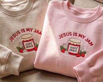 May include: Two crewneck sweatshirts, one beige and one light pink, each featuring an embroidered graphic of a jam jar with the text "JESUS IS MY JAM" and "GOD'S GRACE" on the jar, surrounded by red strawberries and white daisy flowers.