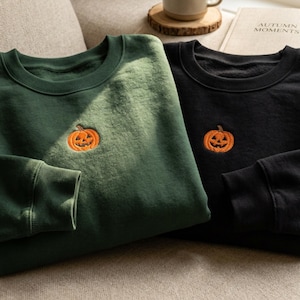 Op de afbeelding: Twee sweaters met ronde hals, één in donker bosgroen en één in zwart, elk voorzien van een klein, centraal oranje geborduurd pompoenontwerp op de borst. Beide kledingstukken hebben lange mouwen met geribbelde boorden en halslijnen.