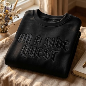 Op de afbeelding: Zwart sweatshirt met de geborduurde tekst "ON A SIDE QUEST" in een gotisch lettertype. Het sweatshirt is netjes opgevouwen op een beige oppervlak, met een raam en een vaas met droogbloemen op de achtergrond.