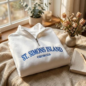 May include: A white quarter-zip sweatshirt featuring blue embroidered text that reads ST. SIMONS ISLAND GEORGIA. The garment has a silver-toned metal zipper pull at the collar and is made of a soft fabric, presented in a folded, clean arrangement.