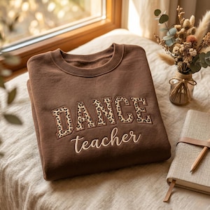 Può includere: Una felpa girocollo marrone scuro con la scritta DANCE in stampatello con motivo leopardato e la parola teacher ricamata in un carattere corsivo color crema chiaro al centro del petto sul davanti.