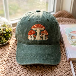Puede incluir: Gorra de béisbol verde con un diseño bordado de setas. Las setas tienen sombreros rojos con lunares blancos y tallos color canela. La gorra está hecha de una tela suave y texturizada y tiene una visera curva.