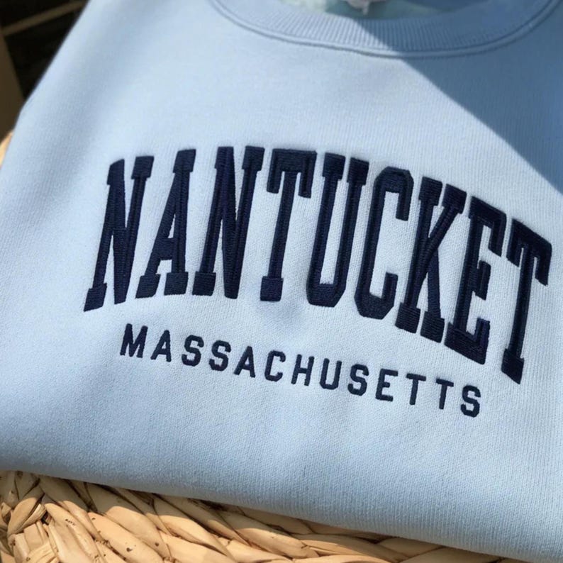 K&ouml;nnte beinhalten: Ein hellblaues Sweatshirt mit Rundhalsausschnitt und dunkelblauer Stickerei, die in einer gro&szlig;en, geschwungenen Schrift NANTUCKET zeigt, wobei das Wort MASSACHUSETTS in einer kleineren, geraden Schrift direkt darunter steht.