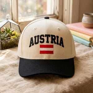 Könnte beinhalten: Eine beige Baseballkappe mit schwarzem Schirm, auf deren Vorderseite das Wort AUSTRIA in schwarzen Großbuchstaben über einem kleinen, rechteckigen rot-weißen österreichischen Flaggensymbol aufgestickt ist.