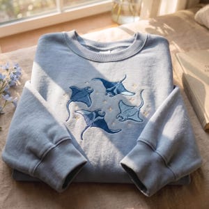 May include: A light blue crewneck sweatshirt featuring an embroidered design of four dark blue manta rays swimming among small, stitched gold stars. The garment has long sleeves with ribbed cuffs and a ribbed neckline, crafted from a soft fabric.
