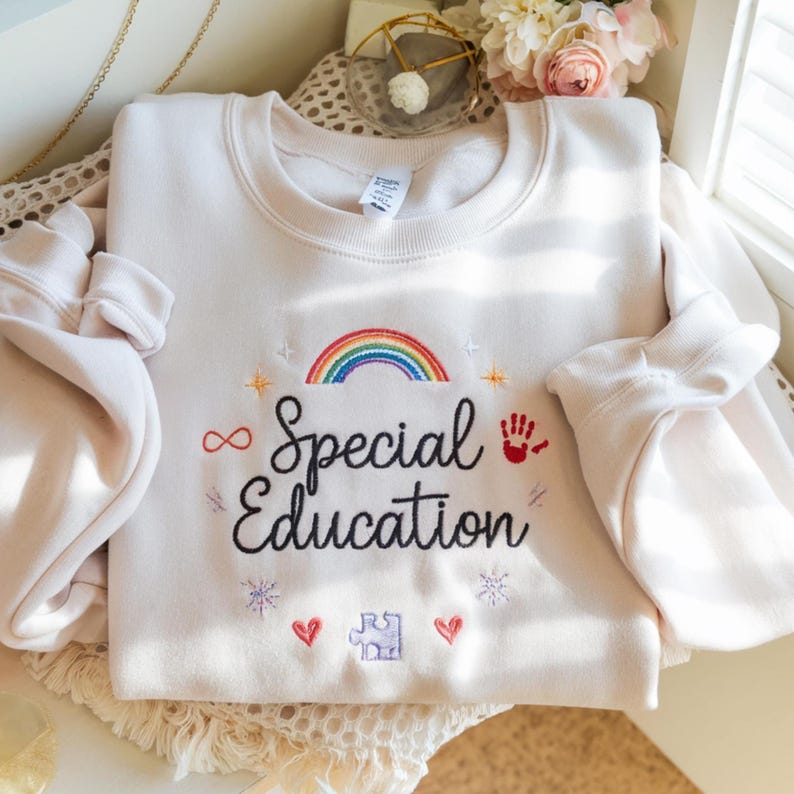 Pode incluir: Camisola creme com arco-&iacute;ris bordado, estrelas, cora&ccedil;&otilde;es, pe&ccedil;a de quebra-cabe&ccedil;a e as palavras "Special Education". O design inclui uma impress&atilde;o de m&atilde;o vermelha e o s&iacute;mbolo do infinito. Uma pe&ccedil;a de roupa confort&aacute;vel e solid&aacute;ria.