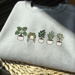 Peut inclure: Un sweat-shirt gris clair avec une broderie de cinq plantes en pot. Les plantes ont des feuilles et des tiges vertes, et sont dans des pots blancs. Le sweat-shirt est plié sur un panier en osier.