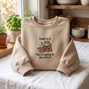 Op de afbeelding: Een beige sweater met ronde hals en een geborduurde afbeelding van een groene kikker in een bruine mantel, die een houten lepel boven een pan houdt. De tekst erboven is THAT'S IT. en de tekst eronder is You're going in the SOUP.