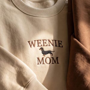 May include: A beige crewneck sweatshirt featuring brown embroidered text that reads WEENIE MOM, with a small embroidered dachshund dog graphic positioned between the two words. The sweatshirt has a ribbed collar and cuffs.