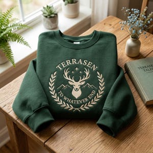 May include: A dark green sweatshirt featuring the word "TERRASEN" above a deer head and mountain design. The text "TO WHATEVER END" is below the design. The design is embroidered in cream. The sweatshirt is on a wooden table.