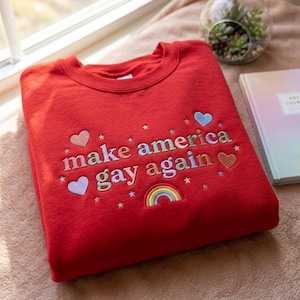 Könnte beinhalten: Ein rotes Sweatshirt mit dem gestickten Schriftzug "make america gay again" in bunten Buchstaben. Das Design umfasst Herzen, Sterne und einen Regenbogen. Das Sweatshirt ist aus weichem Material.