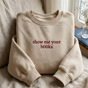May include: A beige sweatshirt with the embroidered phrase "show me your books" in burgundy lettering. The sweatshirt has a crew neck and ribbed cuffs and hem. The garment is folded on a neutral-toned surface.