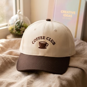 Puede incluir: Gorra de béisbol beige y marrón con la inscripción bordada "COFFEE CLUB" sobre un diseño de taza de café. La gorra tiene una visera curva y un botón en la parte superior.