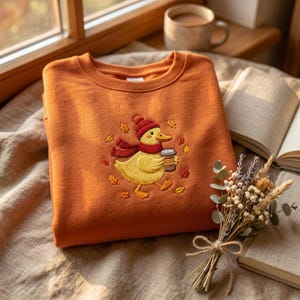 Op de afbeelding: Een roestoranje sweater met ronde hals en een borduursel van een gele eend met een rode gebreide muts en sjaal, die een bruine koffiebeker vasthoudt, omringd door kleine geborduurde herfstblaadjes in de kleuren rood, oranje en geel.
