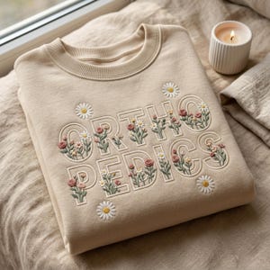 Könnte beinhalten: Beiges Sweatshirt mit dem Wort "ORTHOPEDICS" in Weiß gestickt, verziert mit floralen Designs. Die Blumen umfassen Gänseblümchen und Rosen in Rosa, Grün und Gelb. Eine brennende Kerze ist zu sehen.