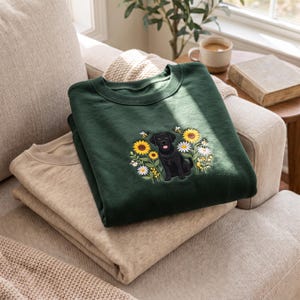 May include: A dark forest green crewneck sweatshirt featuring an embroidered design of a black Labrador retriever sitting among yellow sunflowers, white daisies, and small bees. The sweatshirt is folded and stacked on top of a plain beige sweatshirt.