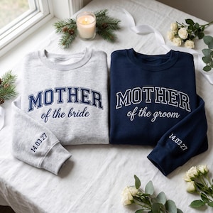 Può includere: Una felpa grigio chiaro con testo blu scuro che recita MOTHER of the bride e una felpa blu scuro con testo bianco che recita MOTHER of the groom. Entrambe le maniche presentano la data ricamata 14.03.27 in colori coordinati.