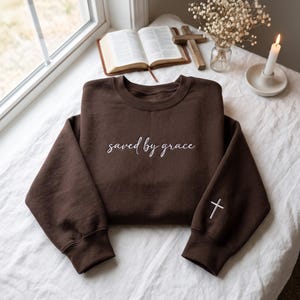 May include: A dark brown crewneck sweatshirt featuring the white embroidered text "saved by grace" across the chest and a small white embroidered cross on the lower portion of the right sleeve, with ribbed cuffs and a ribbed neckline.