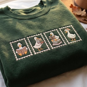 May include: A dark green crewneck sweatshirt featuring four embroidered rectangular patches resembling postage stamps. Each patch depicts a white duck interacting with books, including reading, carrying a stack, sitting on books, and balancing one.