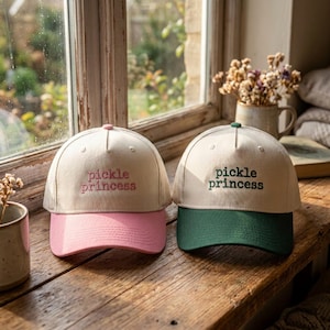 May include: Two baseball caps with off-white crowns and embroidered text reading "pickle princess". The left cap features a pink brim and pink embroidery, while the right cap features a dark green brim and dark green embroidery on the front panel.