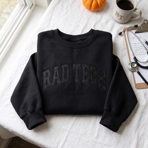 May include: A black long-sleeve crewneck sweatshirt featuring the text RAD TECH embroidered in a matching black thread across the chest in a bold, arched collegiate font. The garment has ribbed cuffs and a ribbed neckline.