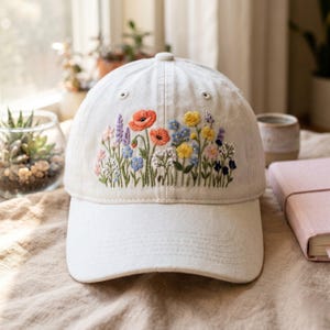 May include: A white baseball cap with a colorful embroidered floral design. The embroidery features red poppies, blue, yellow, and purple wildflowers, and green stems. The cap has a curved brim and a button on top.