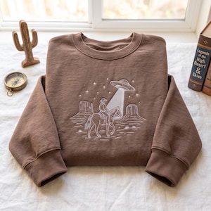 Könnte beinhalten: Ein braunes Sweatshirt mit Rundhalsausschnitt und weißer Linienstickerei, die einen Cowboy auf einem Pferd in einer Wüstenlandschaft mit Mesas, Sternen und einer fliegenden Untertasse zeigt, die einen Lichtstrahl auf das Kleidungsstück wirft.