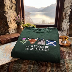 May include: A dark green crewneck sweatshirt featuring embroidered icons of a purple thistle, a brown Highland cow, a grey stone castle, and the blue and white Scottish flag, with the white embroidered text "I'D RATHER BE IN SCOTLAND".