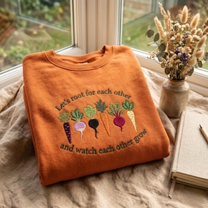 May include: Orange sweatshirt with embroidered vegetables and the text "Let's root for each other and watch each other grow." The design features a purple carrot, a white radish, a black beet, an orange carrot, a red beet, and a yellow parsnip.