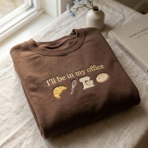 Könnte beinhalten: Ein braunes Sweatshirt mit dem gestickten Schriftzug "I'll be in my office" und Bildern von Croissant, Schneebesen, Mixer und Brot. Das Sweatshirt ist auf einer neutralen Oberfläche gefaltet, mit einem Buch und einer kleinen Vase im Hintergrund.