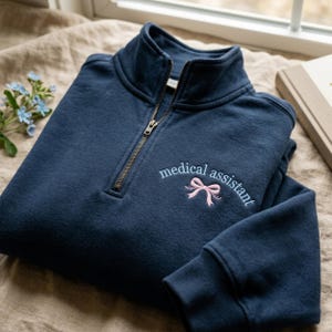 May include: A navy blue quarter-zip sweatshirt with the words "medical assistant" embroidered in light blue, along with a pink bow. The sweatshirt has a ribbed cuff and a brass zipper.