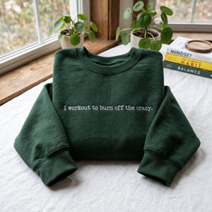May include: A dark forest green crewneck sweatshirt featuring white embroidered text across the chest that reads, i workout to burn off the crazy. The long-sleeved garment has a ribbed collar, cuffs, and hem, presented in a clean, folded style.