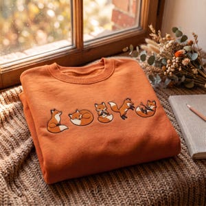 Könnte beinhalten: Ein rostorangefarbenes Sweatshirt mit Rundhalsausschnitt und einer Stickerei von fünf kleinen Füchsen in verschiedenen Posen, darunter sitzend, schlafend, einen Gegenstand haltend, springend und rollend, angeordnet in einer horizontalen Reihe.