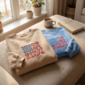 May include: A beige sweatshirt and a light blue sweatshirt, each featuring an embroidered design of blue stars and red seashells arranged in a rectangular pattern. Both garments have long sleeves with ribbed cuffs and a classic crew neckline.