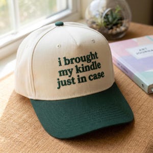 May include: A cream-colored baseball cap with a dark green brim and a dark green embroidered text on the front that reads, "i brought my kindle just in case." The cap features a structured crown with a small button at the top and stitched eyelets.
