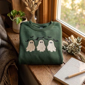 May include: A dark green crewneck sweatshirt featuring an embroidered design of three cream-colored ghosts and several small black bats flying above them. The embroidery is centered on the chest of the garment, which has long sleeves and a ribbed neckline.