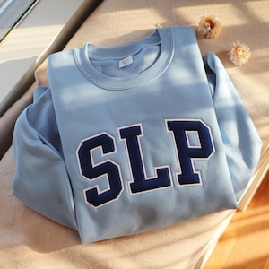 May include: A pale blue sweatshirt featuring the letters "SLP" in navy blue with white edging. The garment is folded on a beige surface. Two small, dried flowers are placed nearby. The sweatshirt is a casual item.