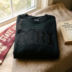 Op de afbeelding: Een zwart sweatshirt met ronde hals en de letters MRI geborduurd in bijpassende zwarte draad op de borst in een lettertype in collegestijl. Het kledingstuk heeft een geribbelde halslijn en lange mouwen, gepresenteerd in een platte vouw.