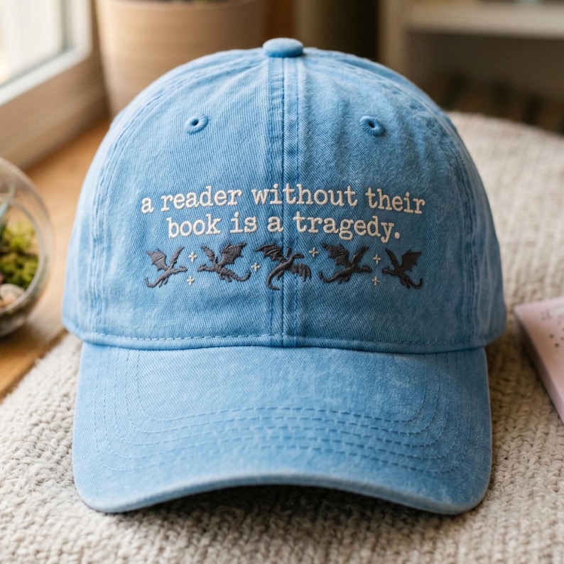 Op de afbeelding: Lichtblauwe baseballcap met geborduurde tekst: "een lezer zonder hun boek is een tragedie." Onder de tekst staan drie geborduurde draken. De cap heeft een gebogen klep en een knoop aan de bovenkant.