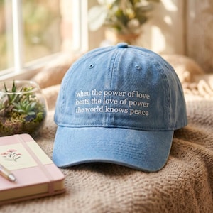 May include: A light blue baseball cap with a distressed finish featuring white embroidered text that reads: when the power of love beats the love of power the world knows peace. The cap has a structured crown and a curved brim with stitching.