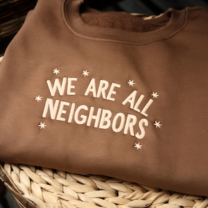 Peut inclure: Un sweat-shirt marron avec l'inscription "WE ARE ALL NEIGHBORS" en blanc cassé, entourée d'étoiles. Le sweat-shirt est plié et posé dans un panier en osier. Le thème général est la communauté et l'unité.