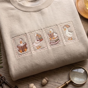 May include: A beige crewneck sweatshirt featuring four embroidered rectangular patches resembling postage stamps. Each patch depicts a white goose interacting with books, including reading, carrying a stack, sitting on a pile, and balancing a book on its head.