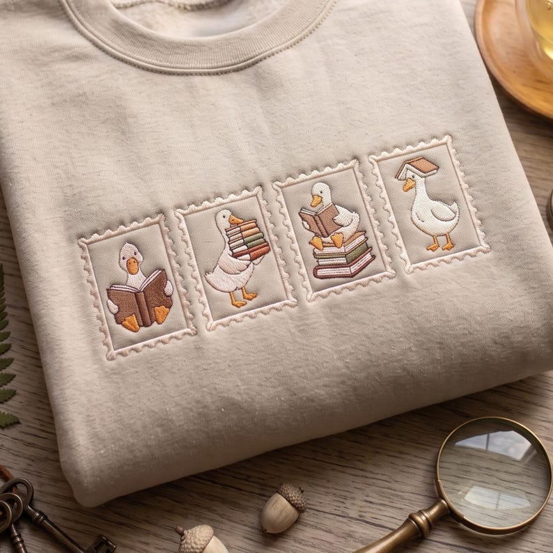 May include: A beige crewneck sweatshirt featuring four embroidered rectangular patches resembling postage stamps. Each patch depicts a white goose interacting with books, including reading, carrying a stack, sitting on a pile, and balancing a book on its head.