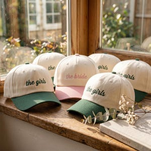 May include: A collection of cream-coloured baseball caps with green brims, featuring embroidered text. Several caps display the words 'the girls' in dark green, while one central cap features the words 'the bride' in light pink embroidery.