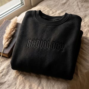以下が含まれることがあります： 胸にゴシック体で「RADIOLOGY」の文字が刺繍された黒いスウェットシャツ。柔らかい素材でできており、クルーネックです。本と装飾用の植物も画像に写っています。