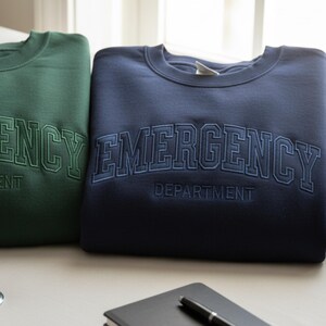 May include: Two crewneck sweatshirts, one in dark green and the other in navy blue. Both feature the word "EMERGENCY" arched across the chest, with "DEPARTMENT" below. The text is embroidered in a matching color.