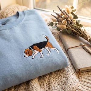 Op de afbeelding: Een lichtblauw sweatshirt met ronde hals en een geborduurde beagle op de borst. De beagle is afgebeeld in een wandelende houding met zwart, bruin en wit haar en draagt een kleine lichtblauwe halsband met een goudkleurig penningetje.