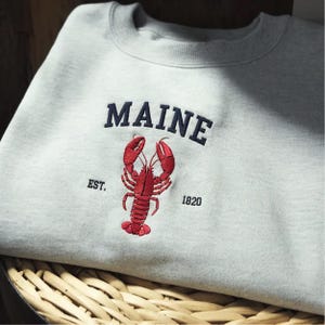 Könnte beinhalten: Ein hellgraues Sweatshirt mit Rundhalsausschnitt und dunkelblau gesticktem Schriftzug MAINE, ergänzt durch EST. und 1820 an den Seiten. Unter dem Text befindet sich eine detaillierte, rote Hummer-Stickerei mittig auf der Brust.